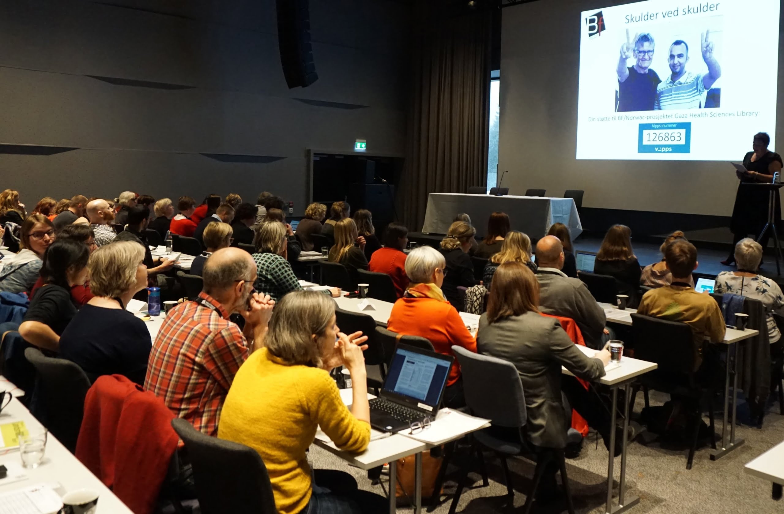 personer sittende ved bord. bilde tatt bakfra, du ser rygger, personer som ser på en scene eller tavle. Foto.