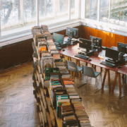 bokhyller i åpent rom ved siden av avlang bord med pcer og skjermer. Vinduer langs ene veggen, utenfor sees et vinterlandskap. foto.