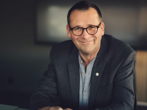 portrett ola eiksund (mann sitter ved bord, smiler, foldede hender foran seg). foto.