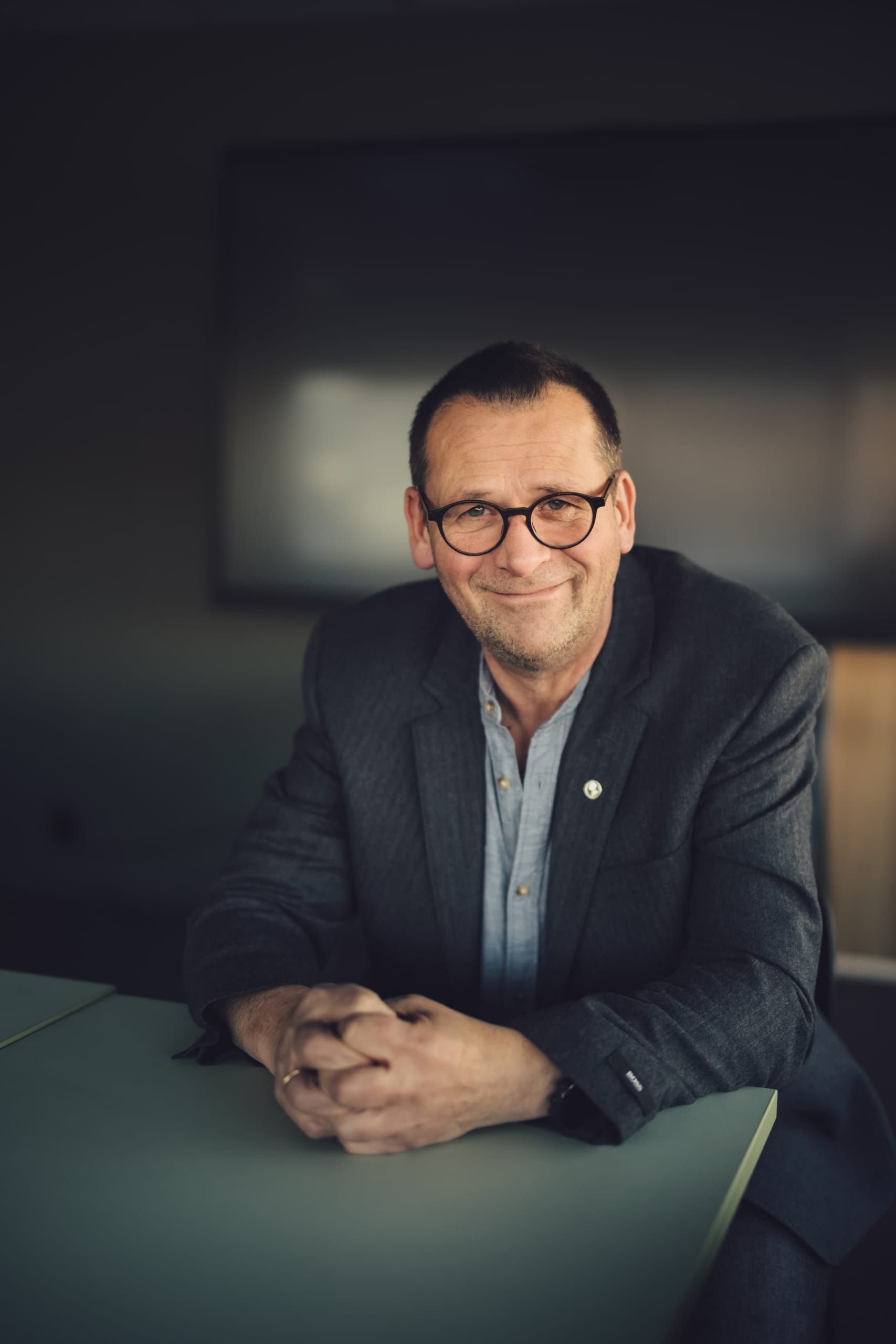 portrett ola eiksund (mann sitter ved bord, smiler, foldede hender foran seg). foto.