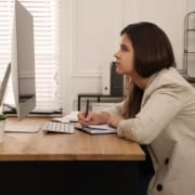 kvinne med krum rygg på stol foran dataskjerm på bord. foto.