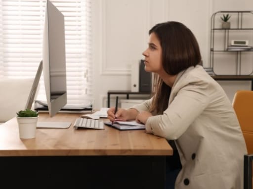 kvinne med krum rygg på stol foran dataskjerm på bord. foto.