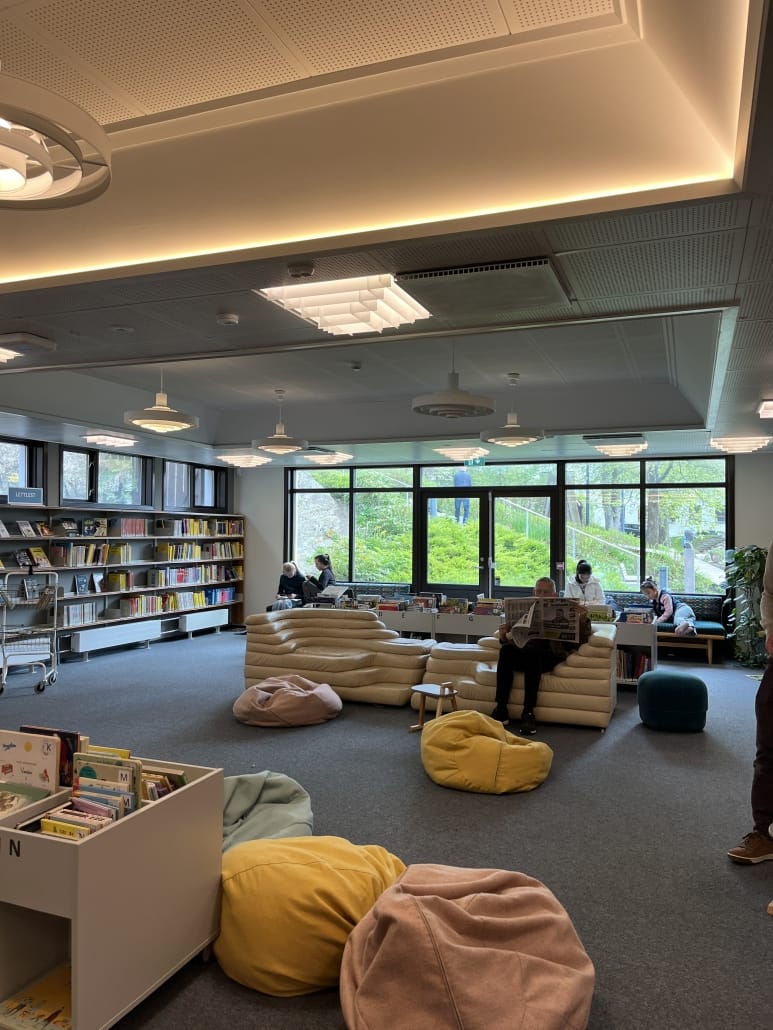 bibliotekrom med sofagruppe der personer leser avisen, i bakgrunnen barn på andre møbler som leser i barnebøker. dempet belysning og tepper på gulv. bøker i hyller på veggene og i kasser rundt som markerer skillene i sonen. foto. 