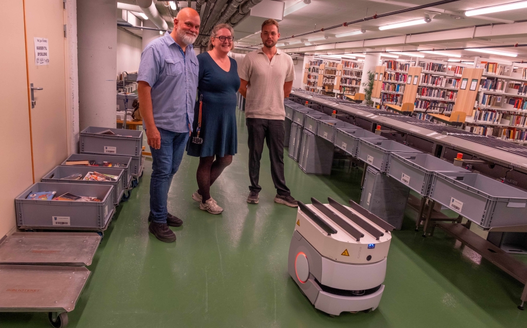tre personer portrettert i et biblioteklokale sammen med en transportrobot. roboten er liten, på hjul og når personene i knehøyde. foto.