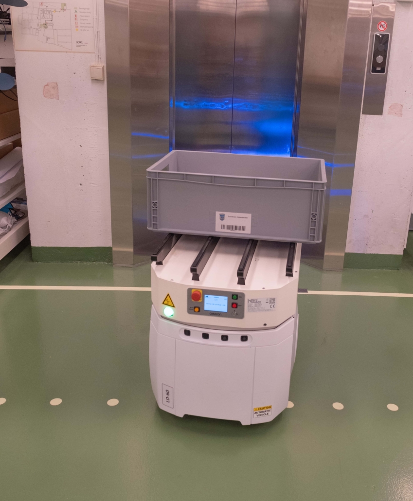 liten kasseformet robot på hjul og skjerm med signaler og knapper og med tom plaskasse plassert på toppen av seg. roboten venter foran en metallfarget heis. foto.