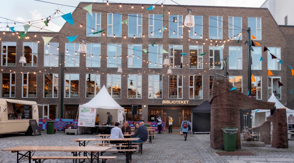 offentlig bygg sett utenfra i flere etasjer, myldrende brosteinsbelagt plass utenfor med benker og besøkende, telt med aktivitetsområde og inngang til biblioteket. foto